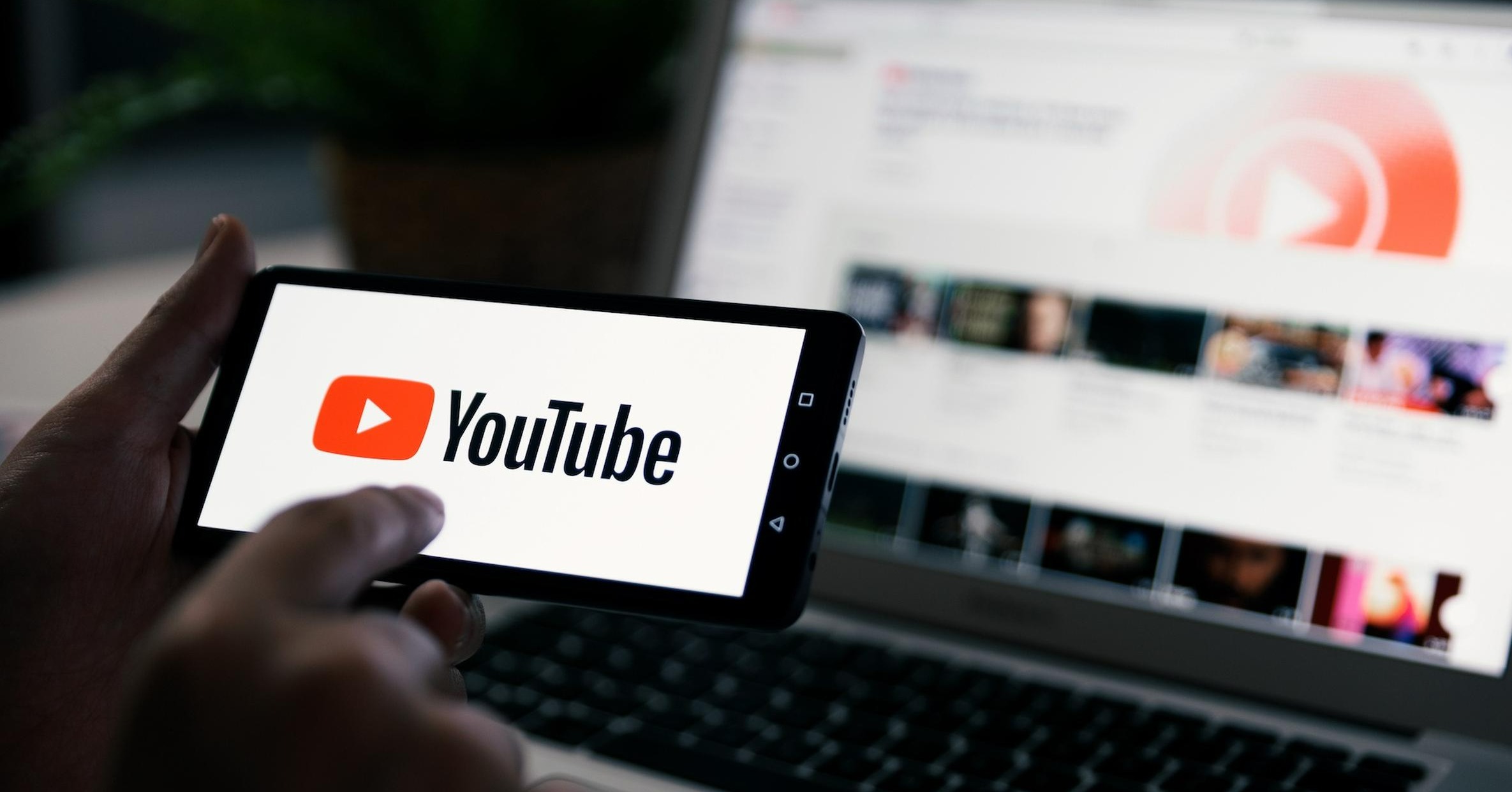 A hand holding a smartphone with the YouTube logo on screen, with a blurred laptop in the background displaying YouTube's interface, illustrating AI content creation for YouTubers