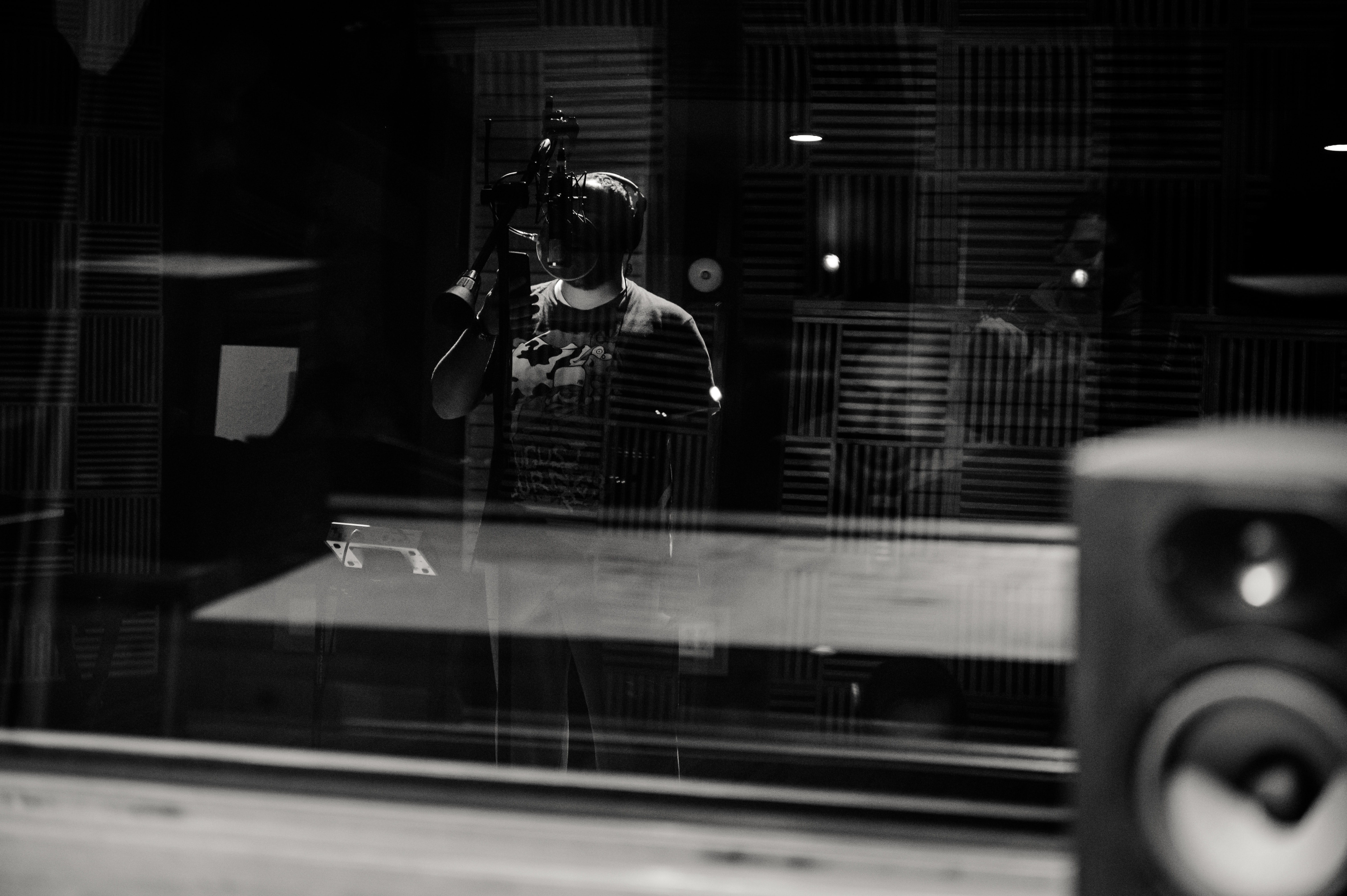 A black and white photo of a person in a recording studio, standing behind a microphone with headphones on, encapsulating the traditional process of voice recording, in contrast to modern voice synthesis methods