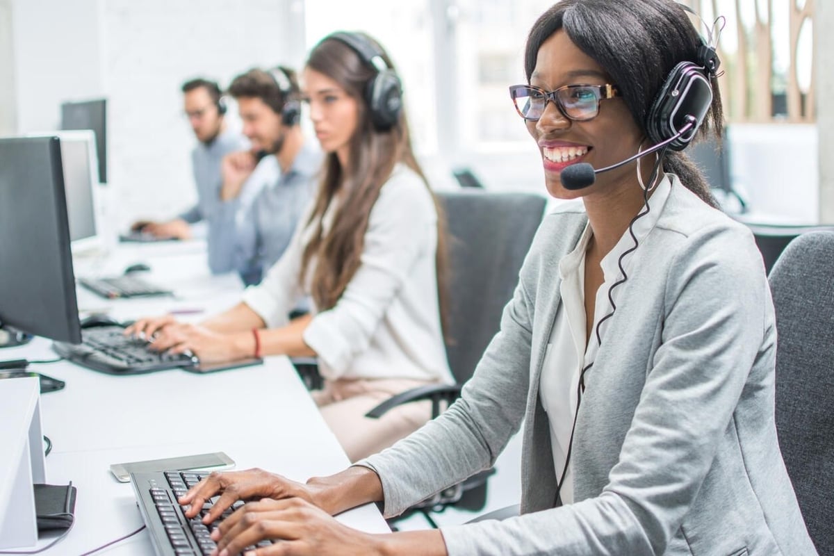 A call center representative with a headset smiling at her computer, exemplifying AI voice synthesis integration for enhanced customer support